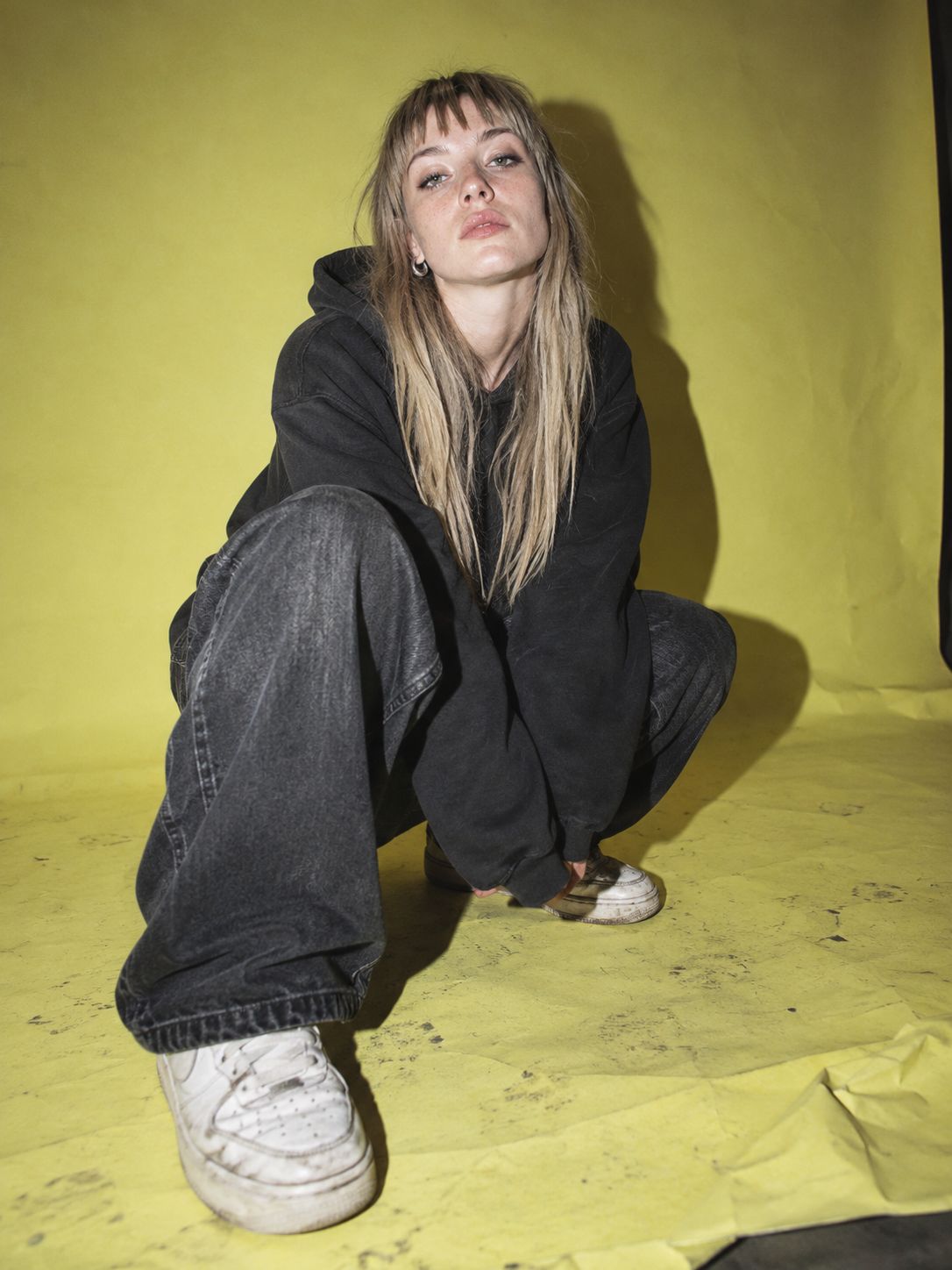 SHAW crouching on a yellow paper backdrop in a black hoodie and wide black jeans.